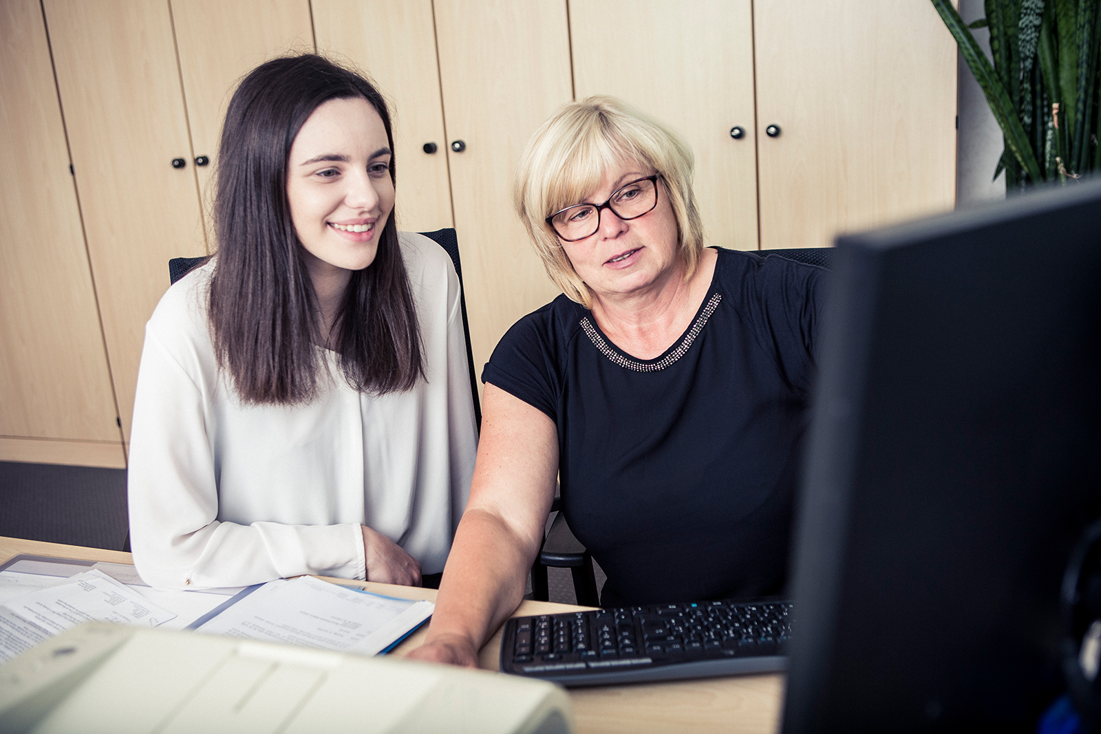 Im Bild sitzen eine ältere Frau und eine Schülerin nebeneinander an einem Schreibtisch. Die ältere Frau zeigt der Schülerin etwas an ihrem Computer. Beide lächeln. Vor ihnen liegen diverse Dokumente. Hinter ihnen ist eine Schrankwand. 