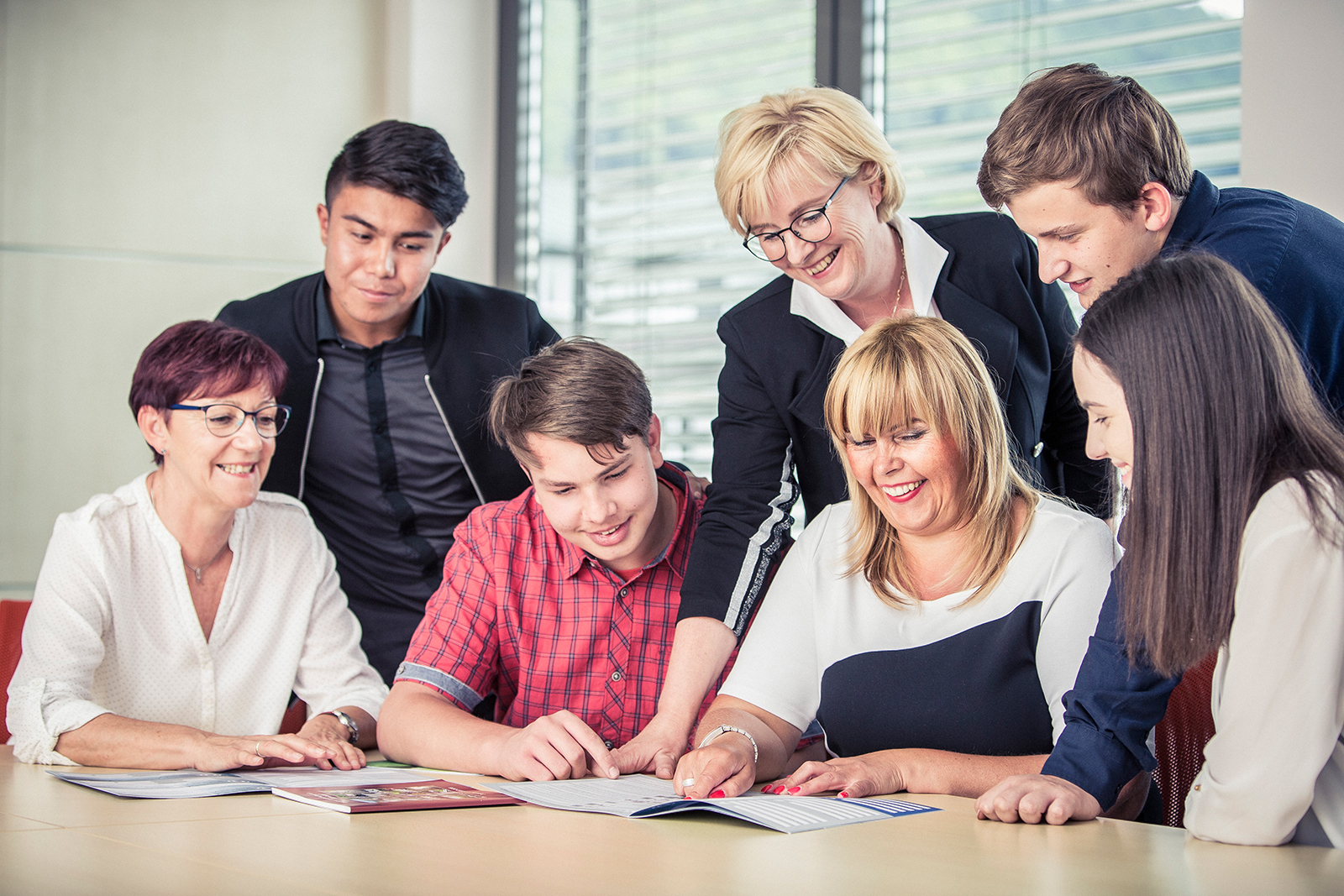 Im Bild sieht man die Prokuristin, Frau Hähnlein, an einem  Tisch sitzen. Um ihr herum sitzen und stehen zwei Lehrerinnen sowie zwei Schüler und eine Schülerin. Frau Hähnlein zeigt auf etwas in einem Prospekt. Alle Personen im Bild lachen freundlich und schauen sich das Prospekt an.