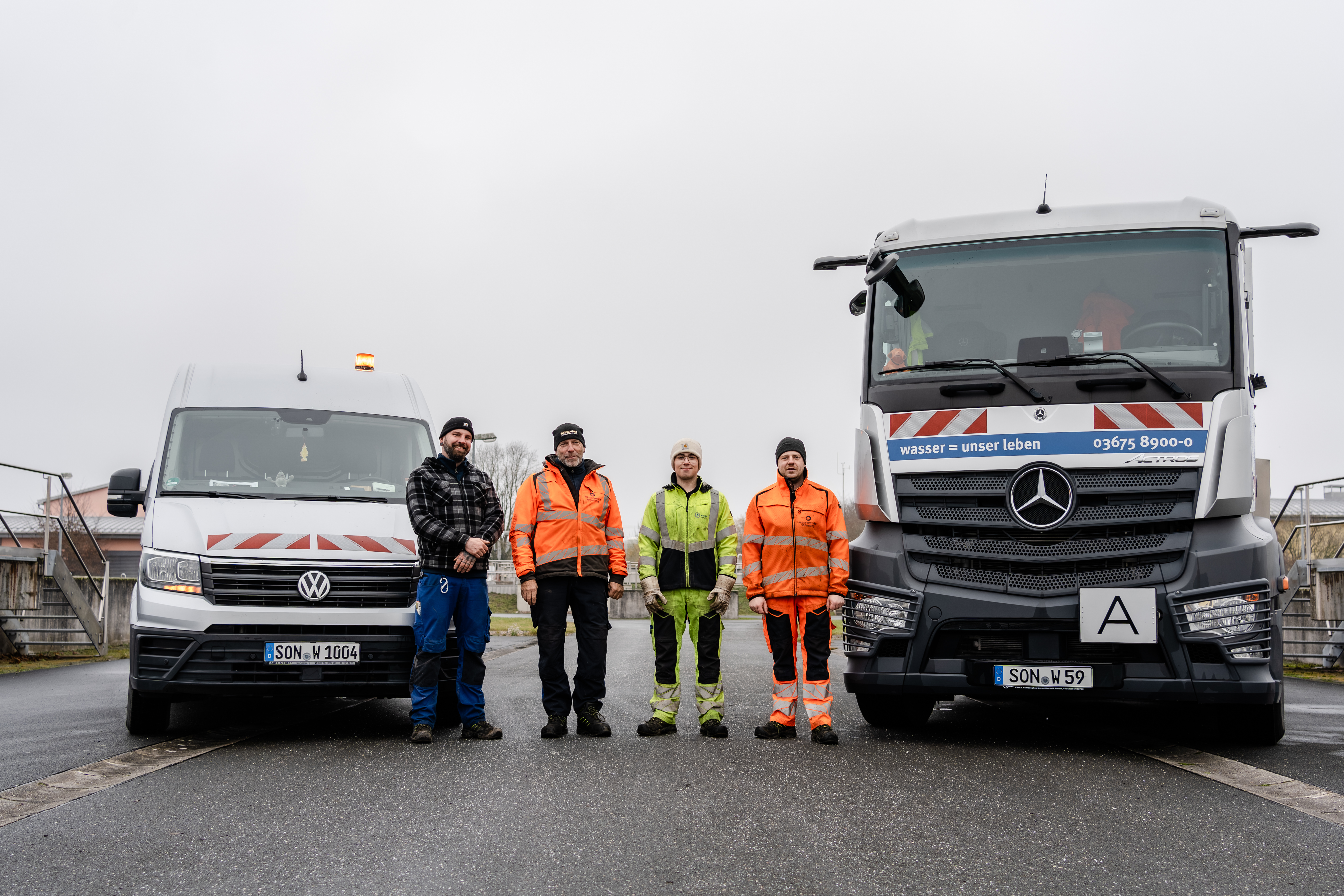 Im Bild sieht man drei Mitarbeiter der Kläranlage. Sie stehen nebeneinander und schauen in die Kamera. Rechts neben ihnen parkt ein VW Bus. Links von ihnen ein steht ein großes Saugfahrzeug für Klärschlamm.