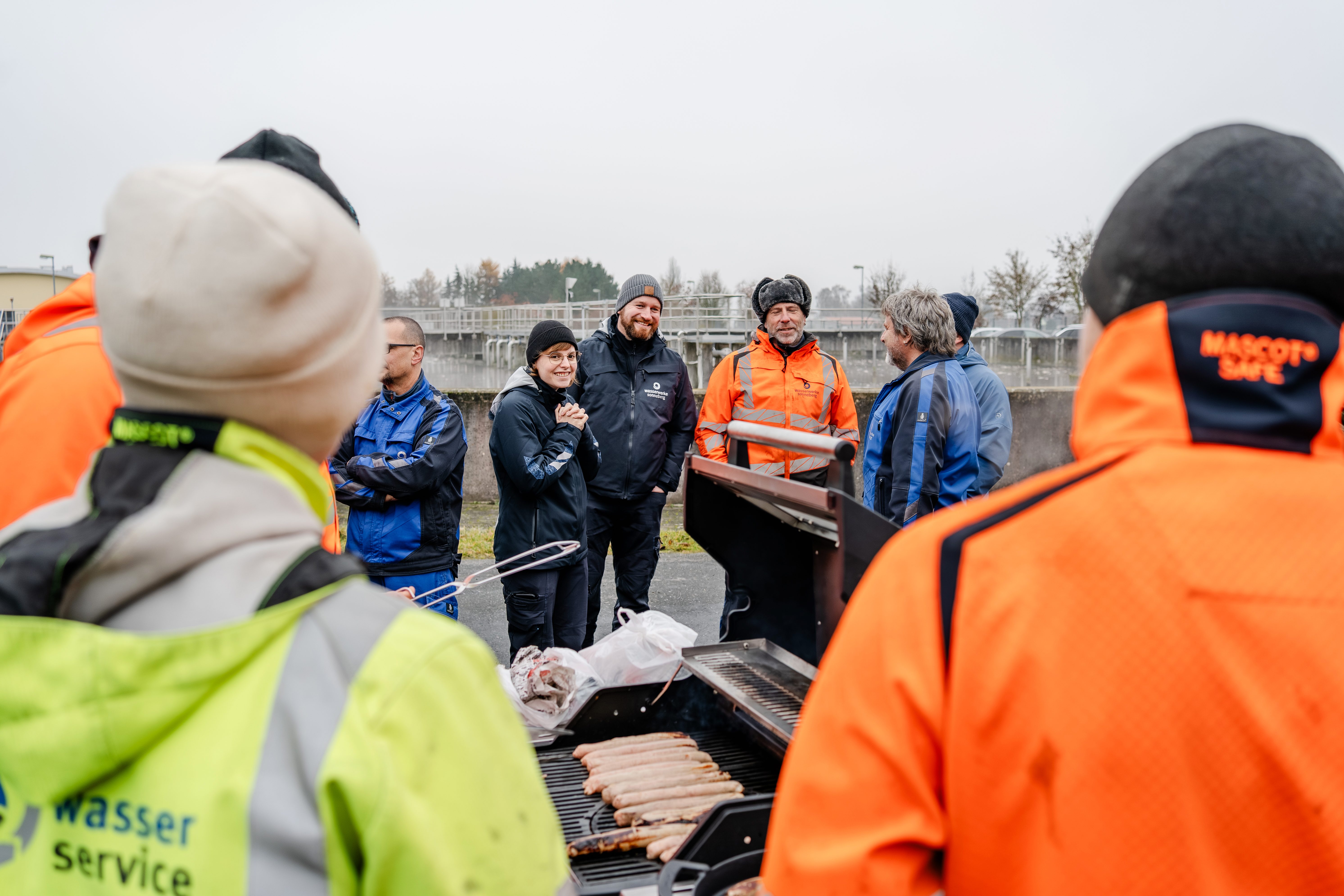 Im Bild sieht man einige Mitarbeiter der Wasserwerke Sonneberg auf dem Gelände der Kläranlage. In der Mitte vom Bild steht ein Grill mit Brautwürsten. Die Mitarbeiter unterhalten sich fröhlich.