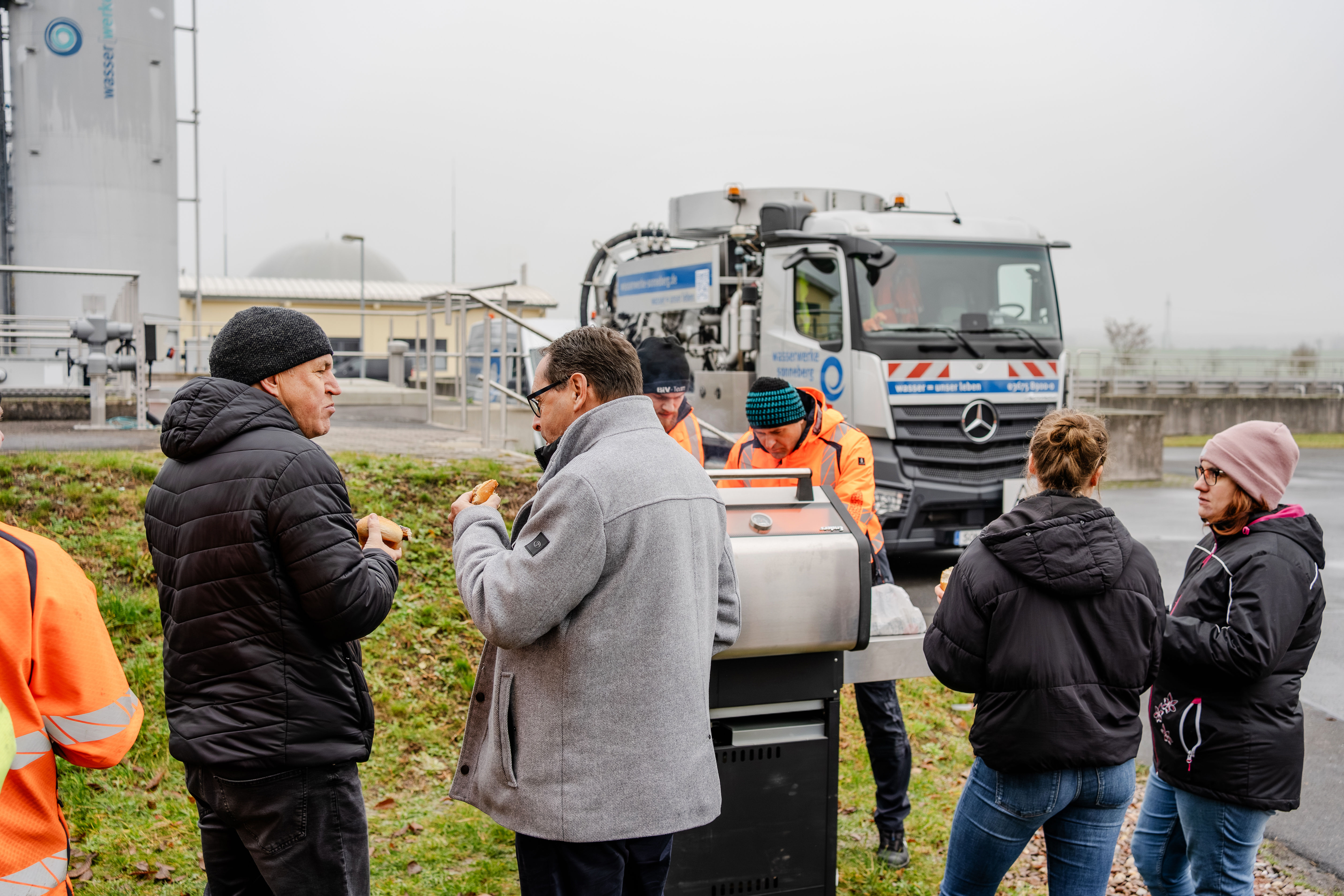 Im Bild sieht man Mitarbeiter und Mitarbeiterinnen sowohl aus den Außenstellen mit Dienstkleidung als auch aus der Verwaltung mit Straßenklamotten. Sie stehen um einen Grill auf dem Gelände der Kläranlage. Zwei von ihnen halten eine Bratwurst in der Hand. Im Hintergrund sieht man ein großes Saugfahrzeug für Klärschlamm.
