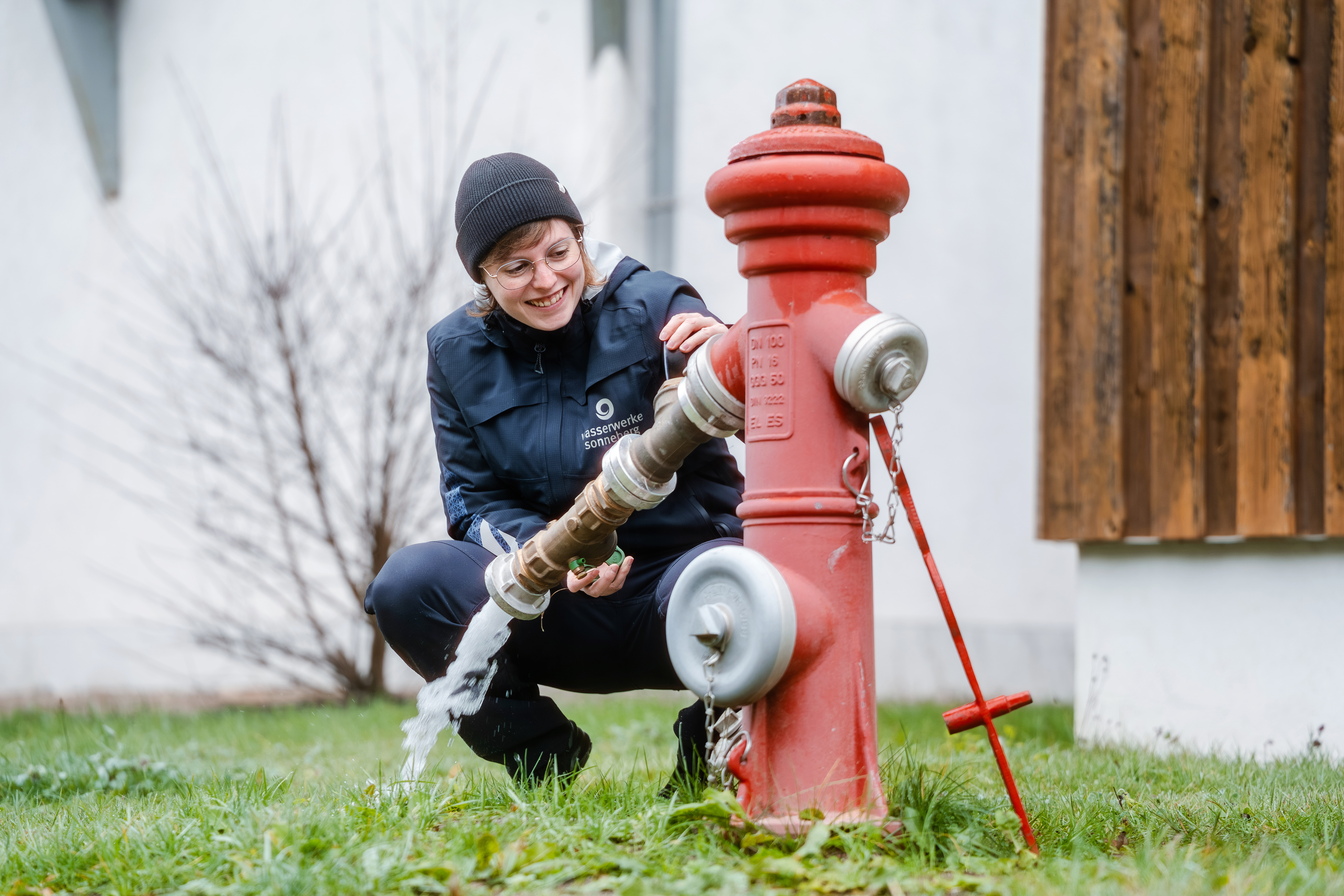 Im Bild sieht man eine Mitarbeiterin aus dem Trinkwasser. Sie hockert neben einem Hydraten, aus dem Wasser fließt. Die Mitarbeiterin prüft den Wasserdruck am Hydranten und lächelt dabei freundlich.